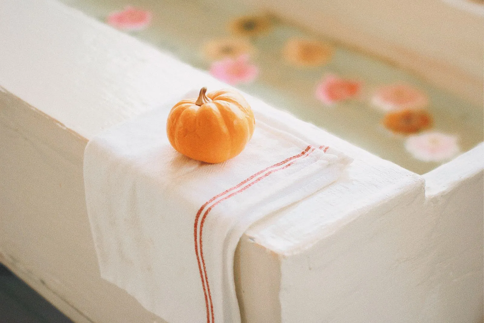 A small pumpkin resting on a towel by a bathtub with floating flowers.
